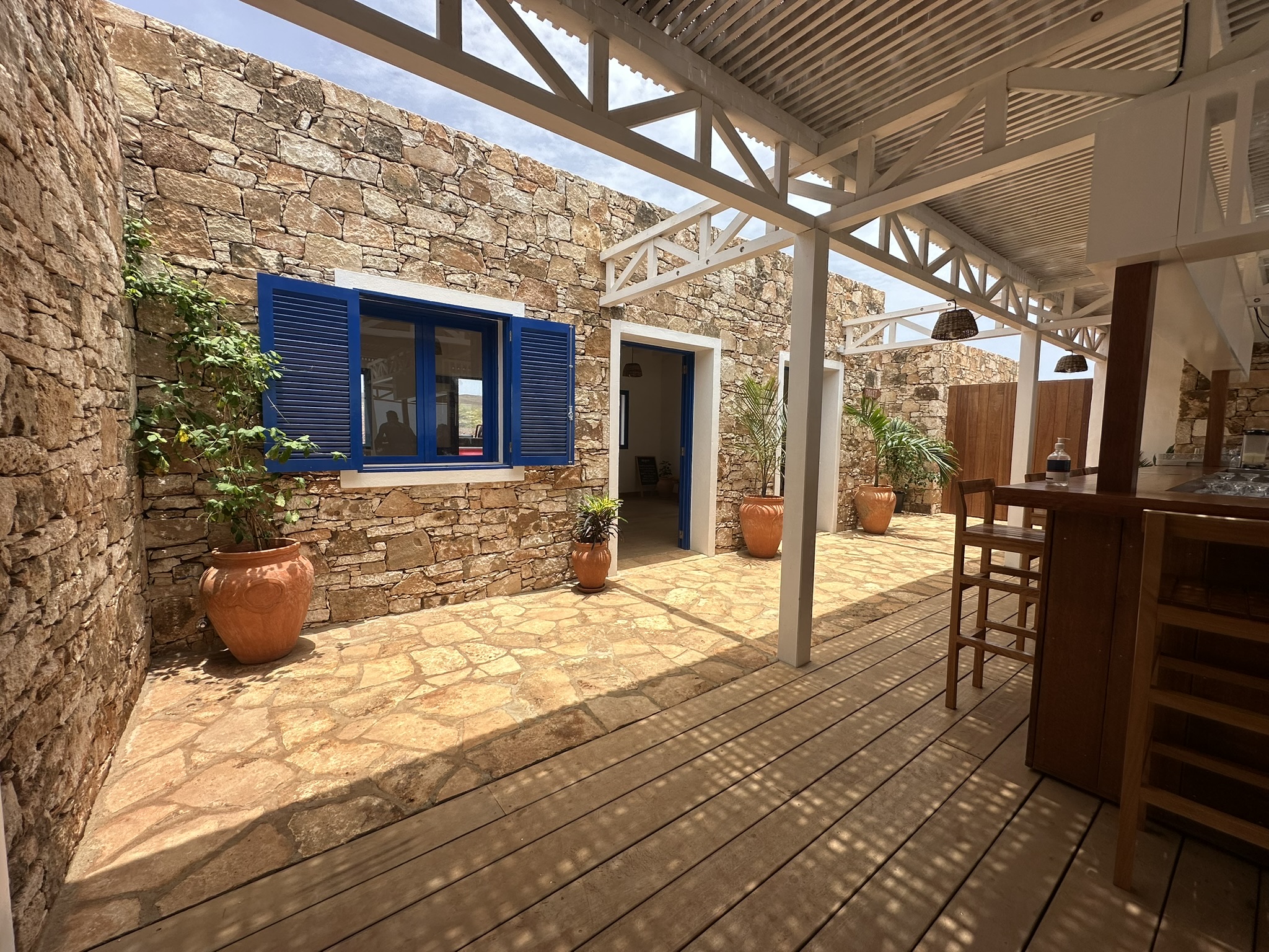 Le patio en pierre — mur de pierre sèche, volets bleus, charpente blanche et jarres en terre cuite